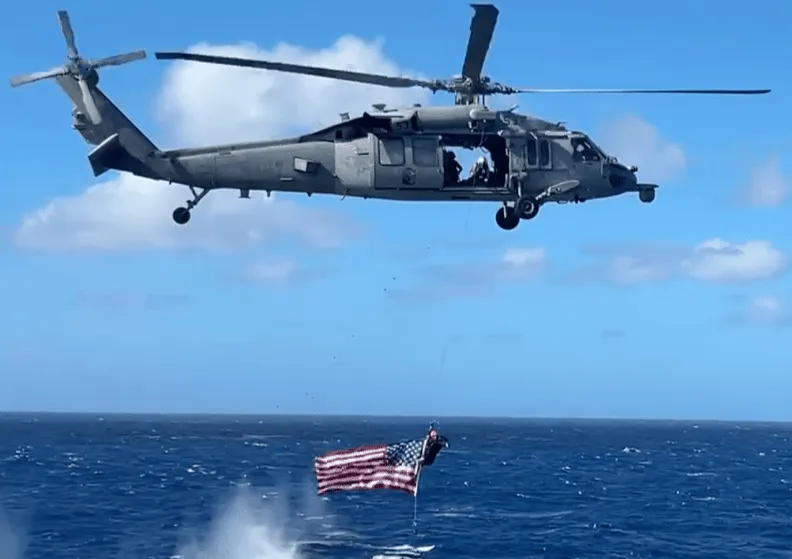 MH-60R Seahawk helicopter hovering over the ocean with a Navy diver below holding an American flag