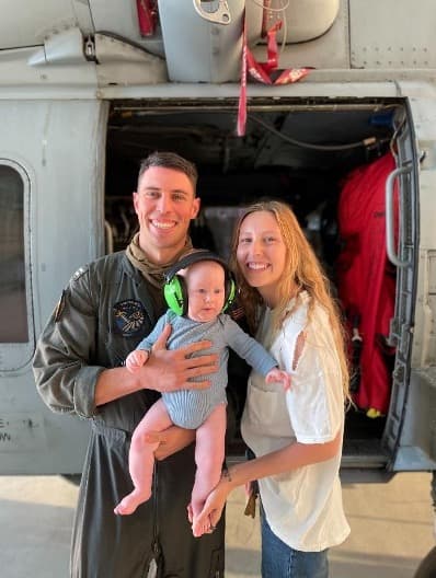 Zach Rodriguez in Navy flight suit holding his baby son with wife Dani in front of an MH-60R Seahawk helicopter